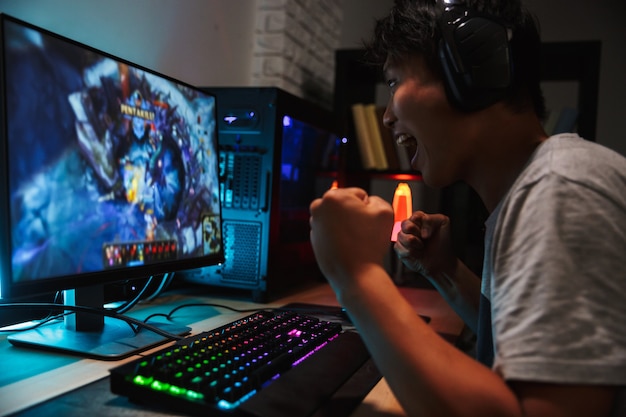 暗い部屋でコンピューターでビデオゲームをプレイし ヘッドフォンを着用し バックライト付きのカラフルなキーボードを使用して興奮しているアジアの10代の ゲーマー少年の肖像画 プレミアム写真