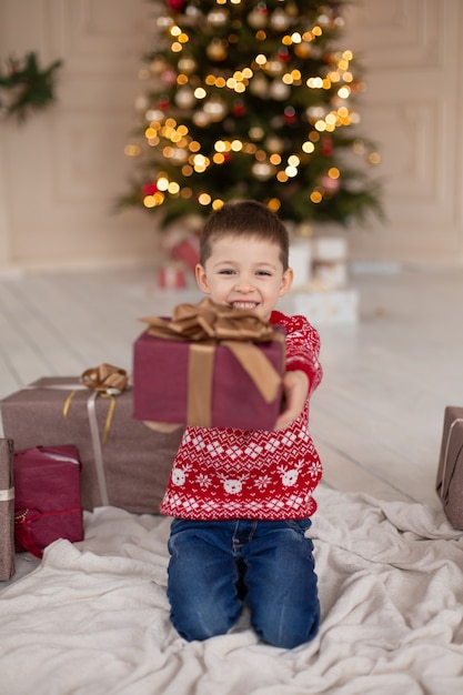 クリスマスツリーの近くにクリスマスプレゼントボックスと赤いニットセーターで幸せな笑顔の小さな男の子の肖像画 幸せな子供時代 クリスマスの話を待っています プレミアム写真