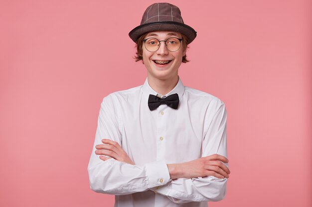 白いシャツ 帽子 黒の蝶ネクタイでうれしそうな面白い若い男の肖像画は ピンクの背景に分離された手を交差させて立って 矯正ブラケットを示して喜んで笑って眼鏡をかけています 無料の写真