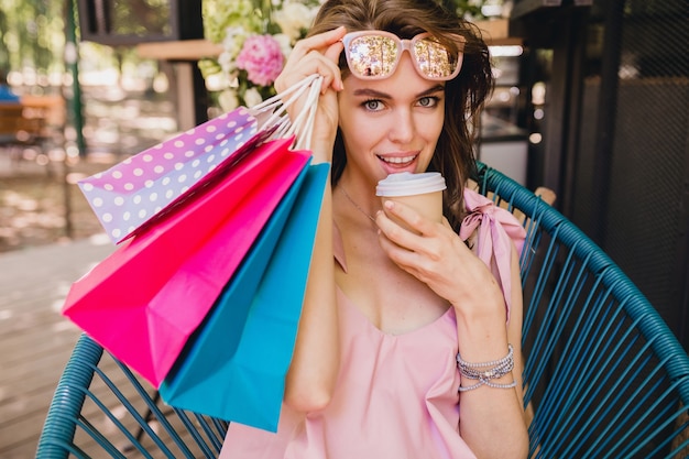 コーヒーを飲みながら買い物袋 夏のファッションの服装 流行に敏感なスタイル ピンクのコットンドレス トレンディなアパレルとカフェに座って興奮した表情で若い笑顔幸せなきれいな女性の肖像画 無料の写真