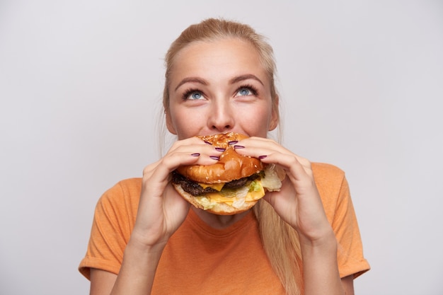 Portrait of pleased young lovely blonde woman with casual hairstyle eating fresh hamburger with great appetite and looking cheerfully upwards, posing over white background Free Photo