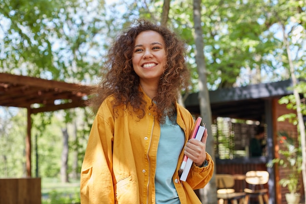 Portrait of smiling young beautiful dark skinned curly student girl on a cafe terrace, holding textbooks, wearing in yellow coat, enjoys the weather. Free Photo