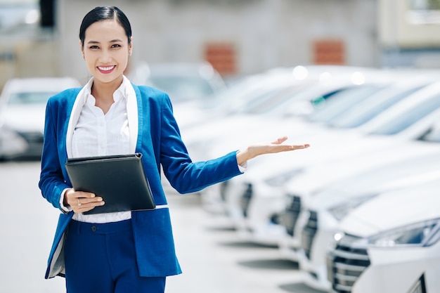 車を選ぶように若い人を招待する革のフォルダーを持つ若い女性のアジアの自動車ディーラーマネージャーを笑顔の肖像画 プレミアム写真