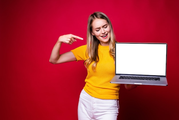 Portrait of smiling young woman pointed on screen of laptop pc computer isolated on red wall. Free Photo