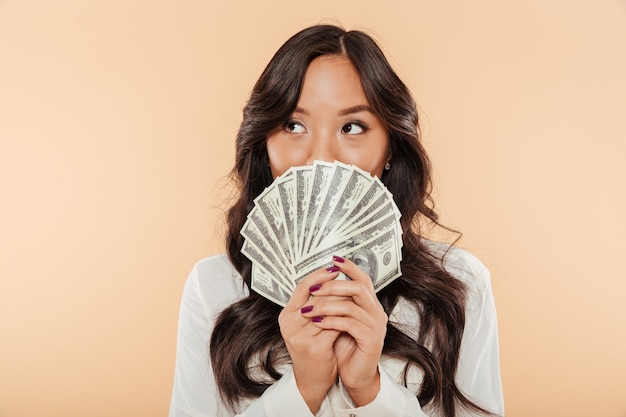 Portrait of successful asian woman covering mouth with fan of 100 dollar bills being satisfied about salary or income posing over beige background Free Photo