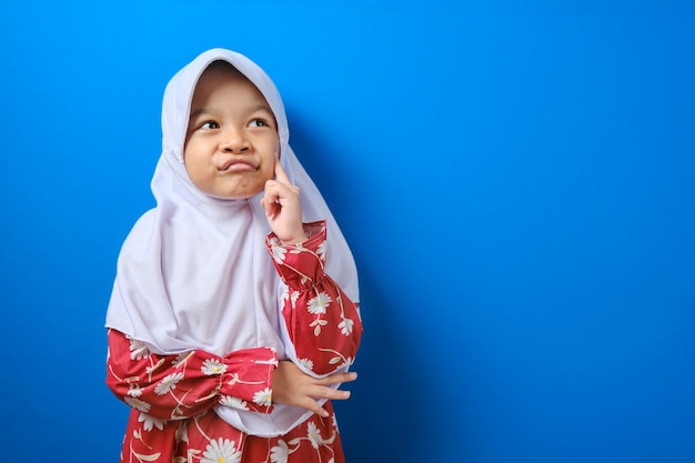 Portrait of young asian muslim girl looked happy, thinking and looking up, having good idea. half body portrait against blue background with copy space Premium Photo