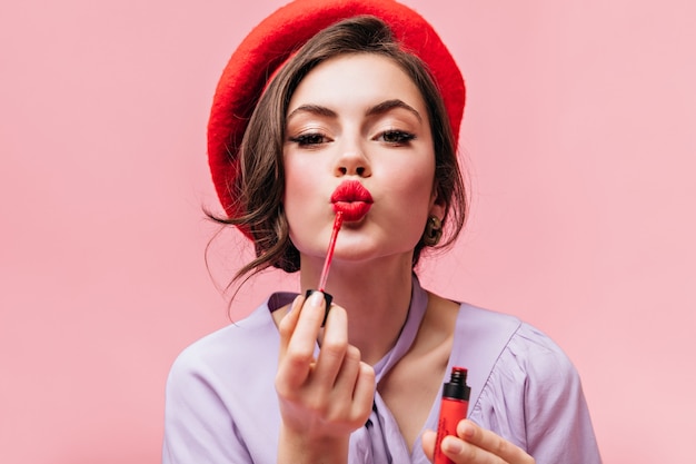 Portrait of young girl in red beret painting her lips with bright lipstick on pink background. Free Photo