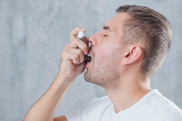 Portrait Of A Young Man Using Asthma Inhaler Against Grey Background 