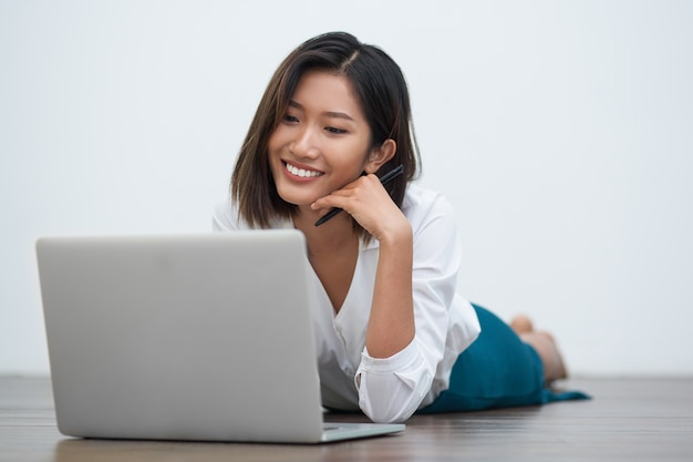 Positive asian woman lying on floor with laptop | Free Photo