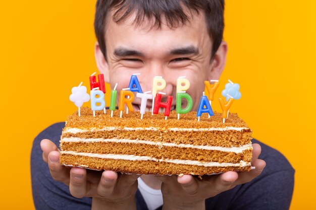 黄色の表面でポーズをとって 帽子と燃えるろうそくと彼の手でケーキを持つポジティブな面白い若いアジア人 プレミアム写真