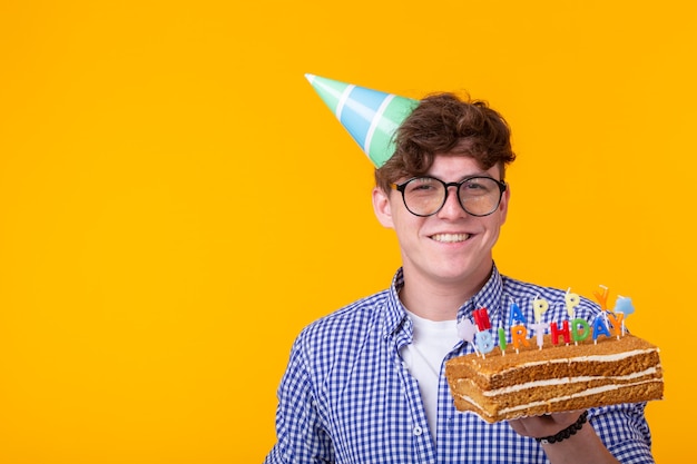 黄色の壁にポーズをとって彼の手に帽子と自家製ケーキを持つポジティブな面白い若い男 プレミアム写真