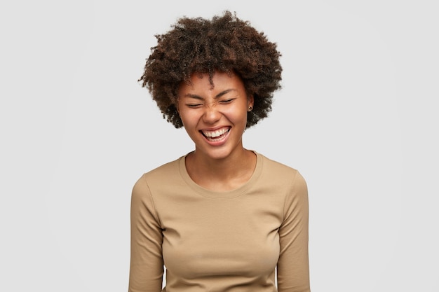 ポジティブな人間の感情の概念 嬉しい表情で幸せな巻き毛の若いアフリカ系アメリカ人女性 笑い 面白いものを笑う 幸せから顔を細くし 白い壁に隔離 無料の写真