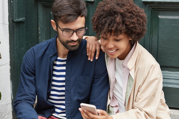 ポジティブな混血カップルが携帯電話で面白いオンラインビデオコンテンツを見て 屋外でポーズをとる 無料の写真