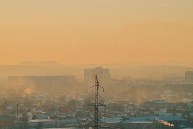 夜明けの都市の送電線 日の出のスモッグの中で都市の建物のシルエット 暖かいオレンジ黄色の空に高電圧のケーブル 日没時の電力産業 市の電源 霧の都市背景 プレミアム写真