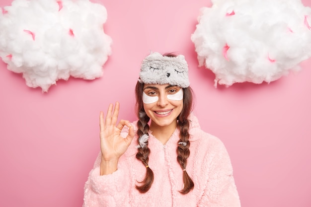 Pretty brunette caucasian woman smiles gently shows okay gesture likes something applies collagen patches to reduce puffiness under eyes dressed in soft nightwear isolated over pink wall Free Photo