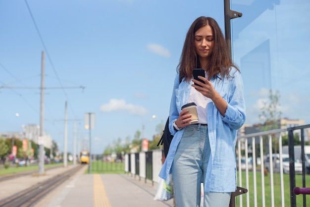 一杯のコーヒーとかわいい女の子は 朝のバスまたは路面電車の路面電車を待ちます アプリを通じてトランスポートを監視するスマートフォンを持つ若い女性 プレミアム写真