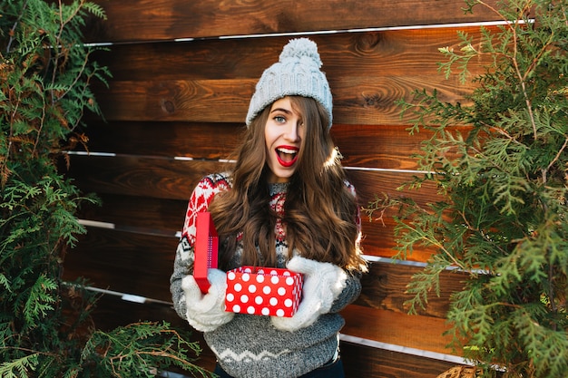 木製の冬の服の長い髪のかわいい女の子 彼女は手袋でクリスマスプレゼントを保持し 驚いて見えます 無料の写真