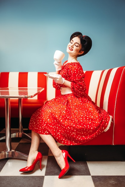メイクアップでかなりピンナップガールはレトロなカフェ 50のアメリカンファッションでコーヒーを飲みます ビンテージスタイルの水玉模様の赤いドレス プレミアム写真