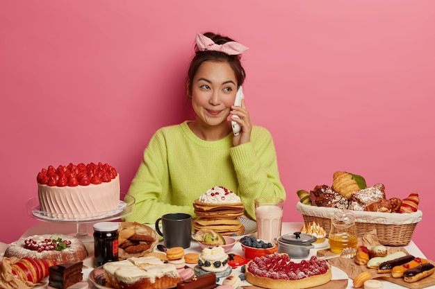 かなりポジティブな韓国の女性は 甘いお菓子やケーキ おいしいおやつを食べ 自分を甘やかし ピンクの背景の上に隔離されて 楽しい電話での会話をしています 無料の写真