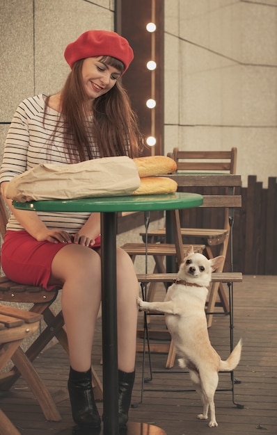 赤いベレー帽のかわいい笑顔の女性は 紙袋にバゲットとカフェのテーブルに座っています プレミアム写真