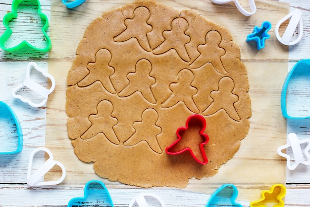 Process of dealing with gingerbread man cookies, use red gingerbread man mold cutting gingerbread dough on baking paper around colorful cookie cutters on white wooden table. top view Free Photo
