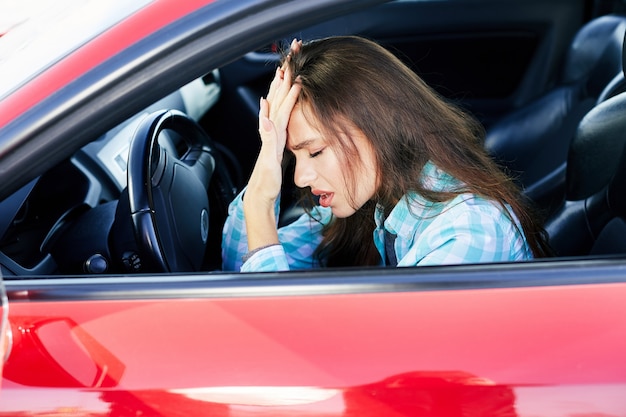 赤い車を運転している神経質な女性のプロフィール 運転中のストレス 目を閉じて手に寄りかかって緊張した女性 交通渋滞 車内のブルネットの女性の頭と肩 プレミアム写真