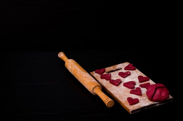 Raw red heart-shaped cookies and dough on wooden board with flour, wooden rolling pin. black . preparing for valentine's day Premium Photo
