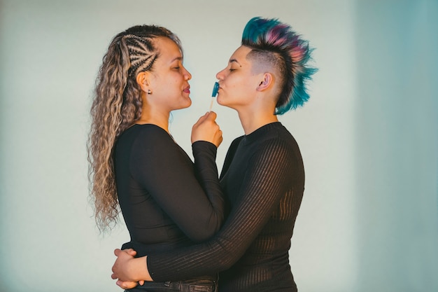 本当のレズビアンのカップル 青いモヒカン髪型の美しいパンク女性にキスするハート型のロリポップで遊ぶ民族の同性愛者のガールフレンド プレミアム写真