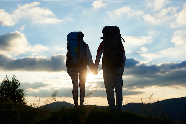 後ろ姿のバックパックに手をつなぐ二人は 雲と空の下で山の明るい夕日を楽しんでいます 山でロマンチックな夜 プレミアム写真