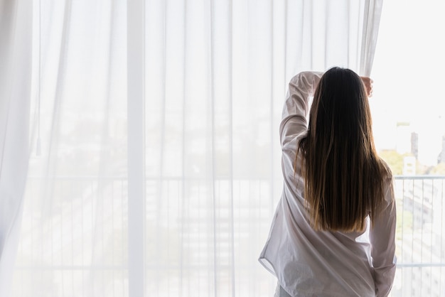 Retrovisor de uma jovem abrindo as cortinas pela manhã Premium Photo