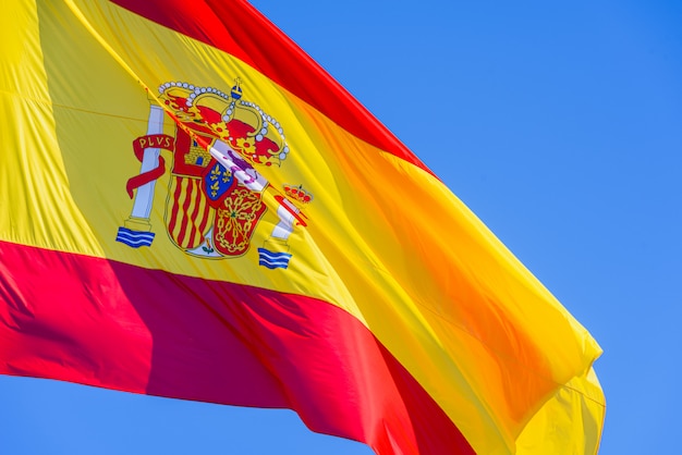 プレミアム写真 青い空を背景に分離された風になびかせてロイヤルシールドと赤と黄色のスペイン国旗
