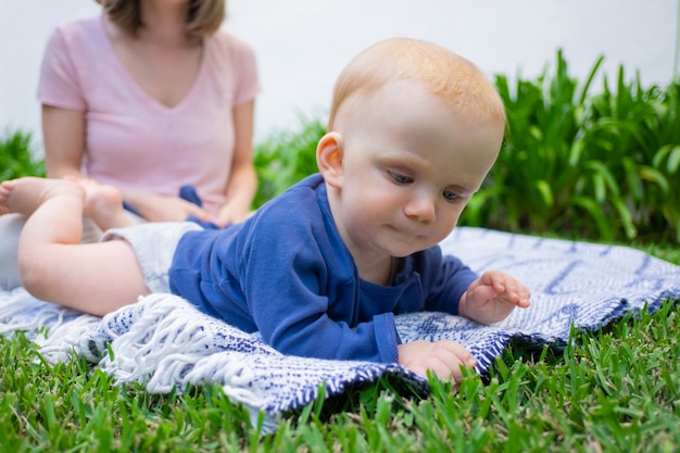 赤毛の女の赤ちゃんは格子縞の上に横たわって 興味を持って草に触れ よそ見します 庭のポートレート クローズアップ 座っている若いお母さん 夏の家族の時間 晴れた日 新鮮な空気の概念 無料の写真