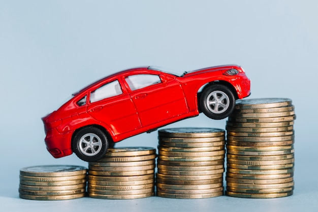 Red small car riding over the increasing coin stack against blue background