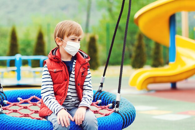 一人でブランコに座っているフェイスマスクの悲しい少年 空の遊び場で退屈した子供 コロナウイルス検疫 プレミアム写真