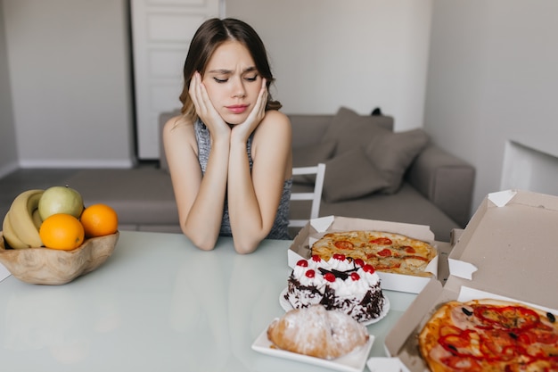 ダイエット中にケーキを見ている悲しい巻き毛の女性 果物とピザでポーズをとる金髪のゴージャスな女性モデル 無料の写真
