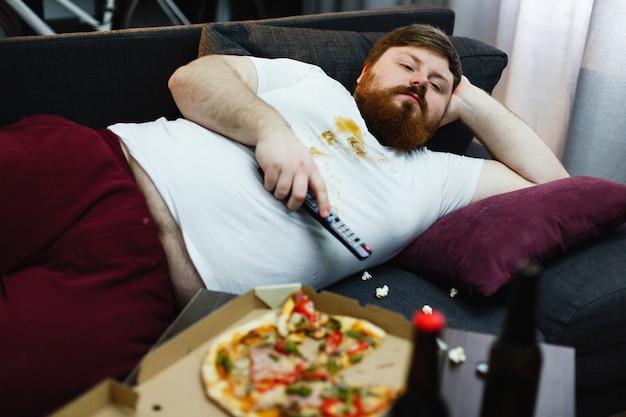 Sad fat man lies on the sofa and watches TV Free Photo