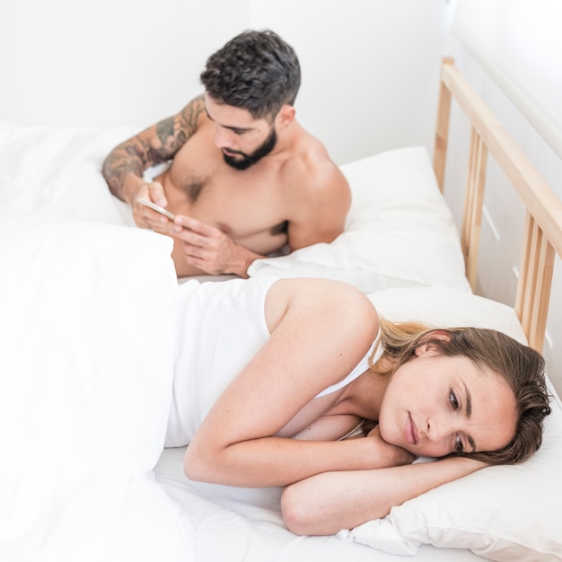 Sad woman lying on bed in front of her husband using mobile phone Free Photo