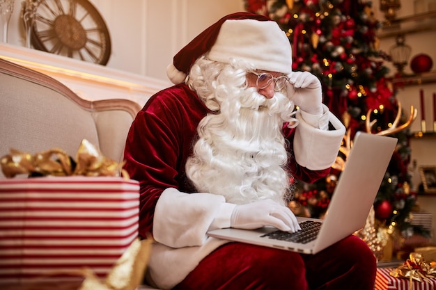 Premium Photo Santa Claus Sitting At His Home And Reading Email On Laptop With Christmas Requesting Or Wish List Near The Fireplace And Tree With Gifts