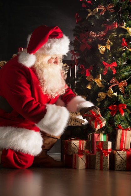 Santa putting gifts under Christmas tree in dark room Photo | Premium ...