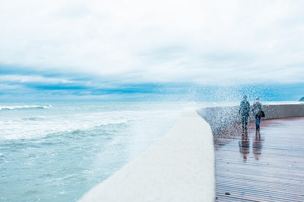 Premium Photo Scary Stormy Waves With Big Sea Wave Splashing Over Pier Road At Cloudy Autumn Rainy Day High Wave Is Breaking On The Pier Two People Are Walking On The