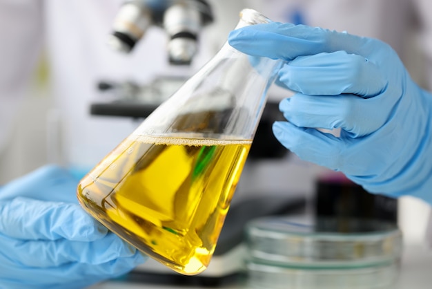  Scientist chemist holding glass flask with yellow liquid in front of microscope closeup quality