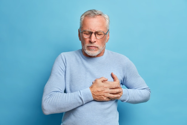 Senior man in spectacles presses hand to chest has heart attack suffers from unbearable pain closes eyes wears optical glasses poses against blue wall Free Photo