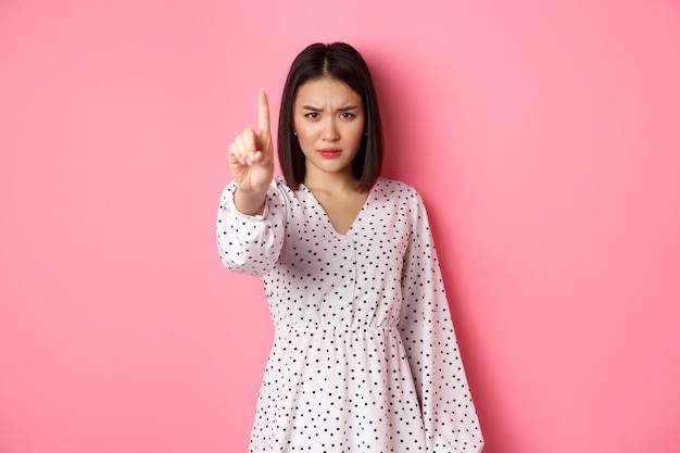 Serious and angry asian woman telling to stop, frowning and showing finger in disapproval, prohibit something, standing in dress against pink background. Free Photo
