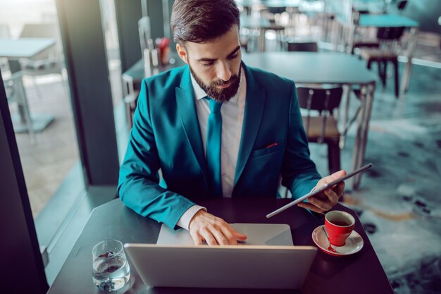 Premium Photo Serious Caucasian Bearded Businessman In Suit Using Tablet And Laptop In The Same Time While Sitting In Cafe On Desk Coffee It Takes Time For Success Just Be Patient