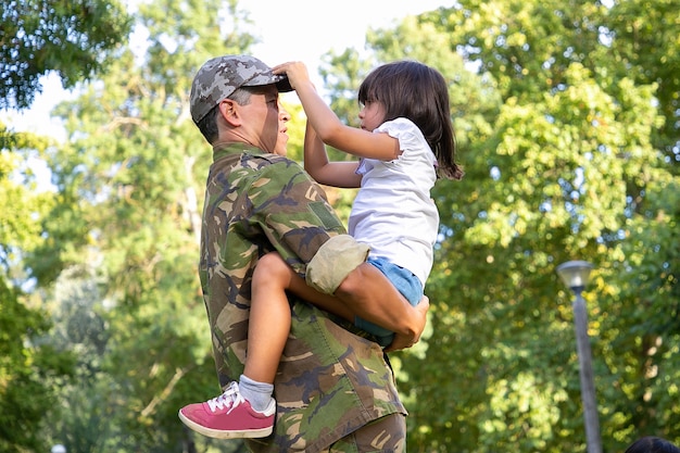 軍服を着た真面目なお父さんが娘を手に持って 彼女を見て 屋外に立っています 父の帽子に触れる集中した小さなかわいい女の子 家族の再会 父性 週末のコンセプト 無料の写真