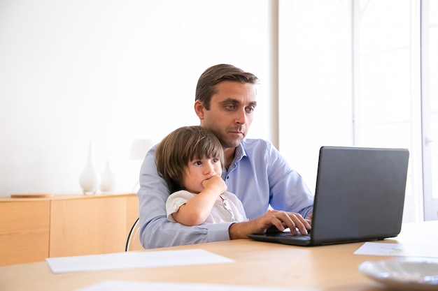 ノートパソコンでインターネットを閲覧し かわいい息子を抱いて真面目な父親 シャツを着て 子供と一緒にテーブルに座って 画面を見て 働いている白人の中年男性 父権と家の概念 無料の写真