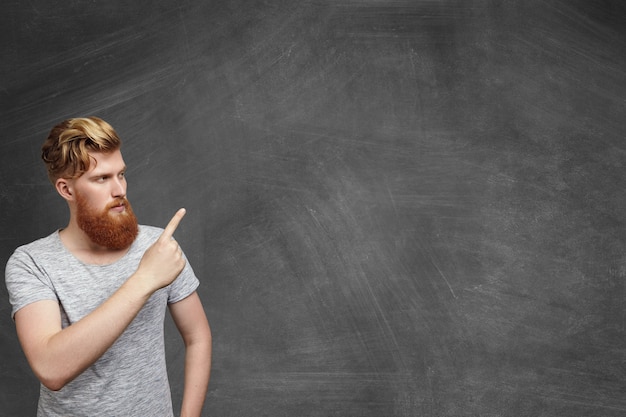 Serious redhead bearded caucasian hipster student dressed in gray t-shirt standing in classroom pointing at blank copy space wall with his finger, showing something on it. Free Photo