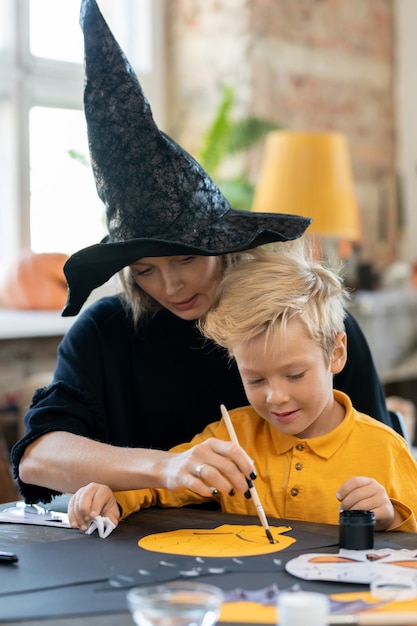 魔女の帽子をかぶった真面目な若い母親が息子と膝をついて座って ハロウィーンの装飾のためにスケルトンを描くのを手伝っています プレミアム写真