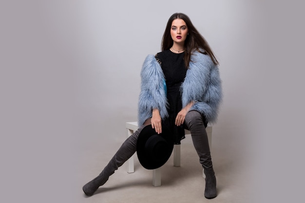 Sexy brunette woman in trendy blue fur jacket and velvet thight high boots posing on grey wall.