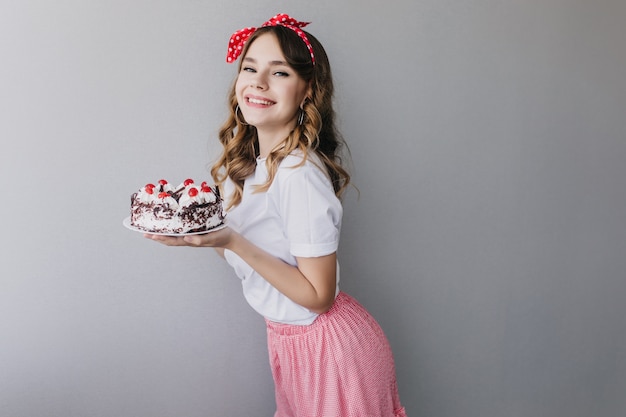 甘いパイを保持している赤いリボンを持つ形の良い女性モデル バースデーケーキを持っている陽気な巻き毛の女性の屋内ショット 無料の写真